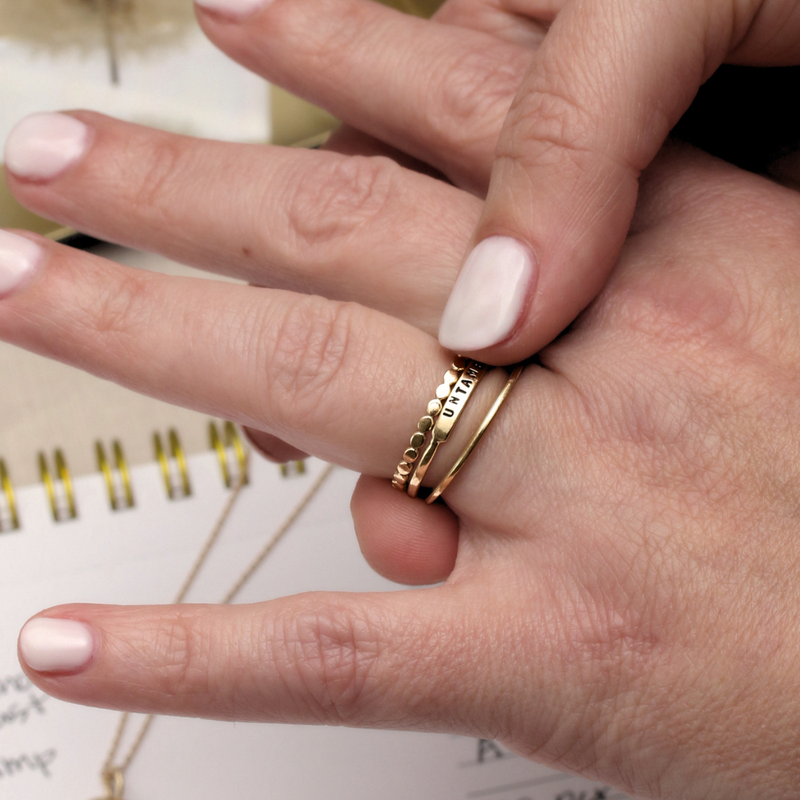  Two hands resting together over an open spiral notebook, wearing a stacked gold ring set with "UNTAMED" stamped on the narrow band, studio details visible in the soft background.